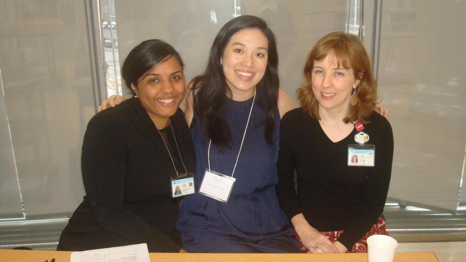 3 female doctors posing for picture seated at wooden table with windows behind them