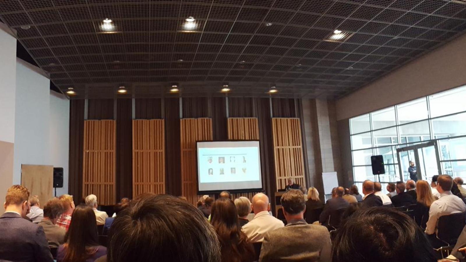 Large conference room full of seated people with presentation board in front and full wall of windows on right side