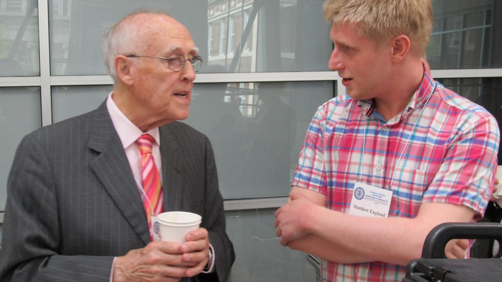 One man in dark suit holding coffee speaking to younger man in plaid shirt.