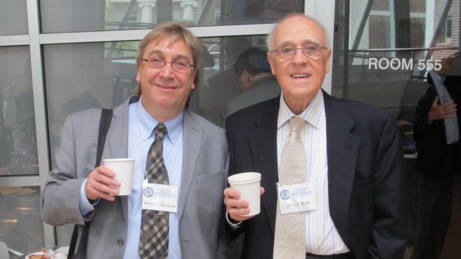 Two men in suits posing for picture with coffee cups in right hand
