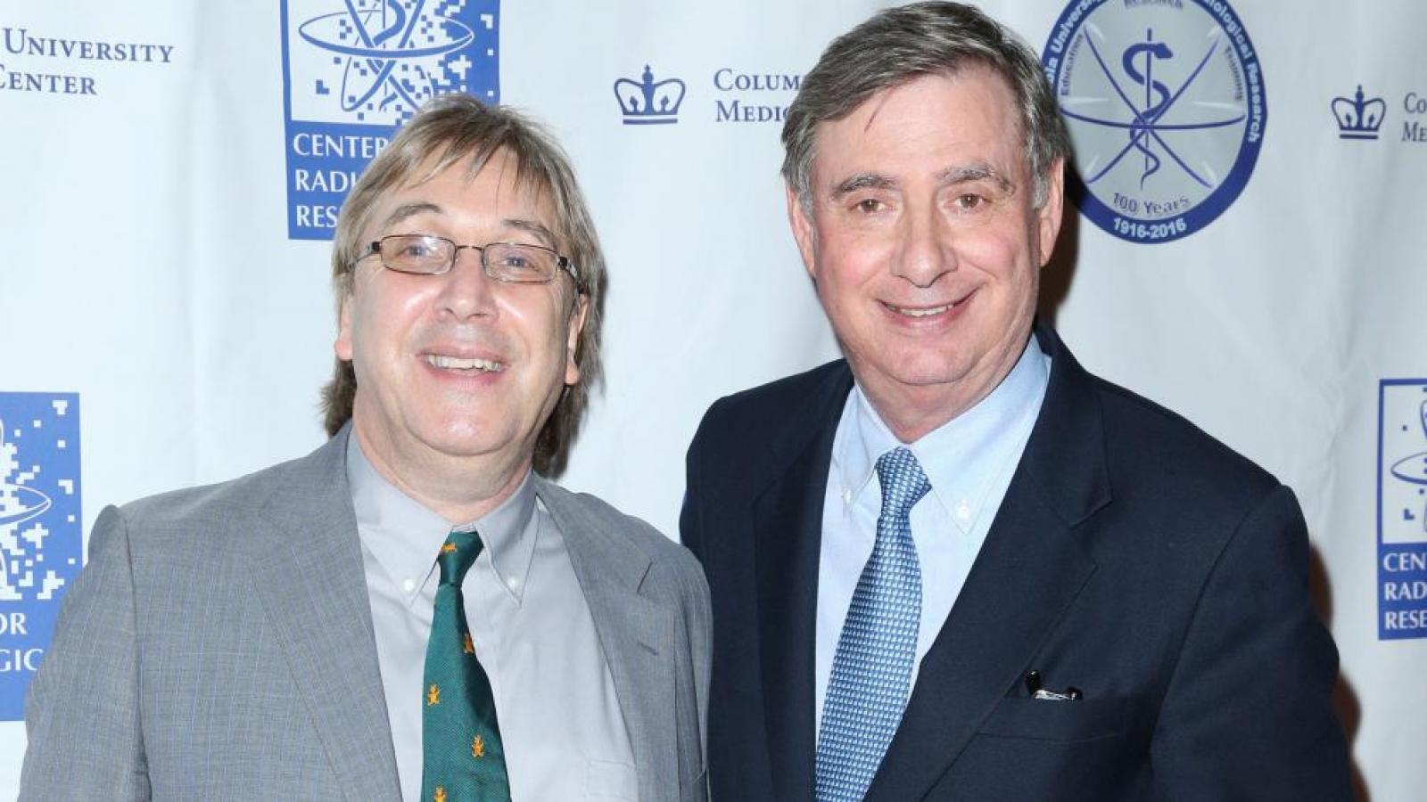 Man in gray suit with man in navy blue suit posing for picture in from of backdrop with logos on it