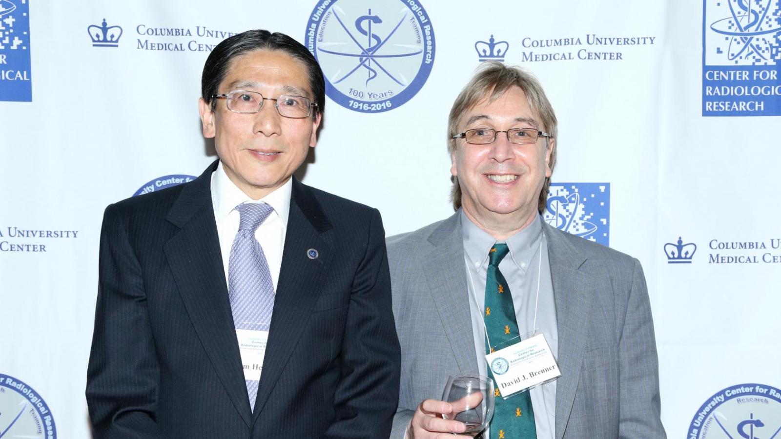 Man in dark blue suit with man in gray suit holding wine glass posing for picture in front of backdrop