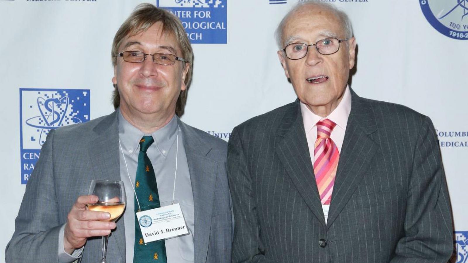 Two men in suits posing for picture in front of back drop with logos
