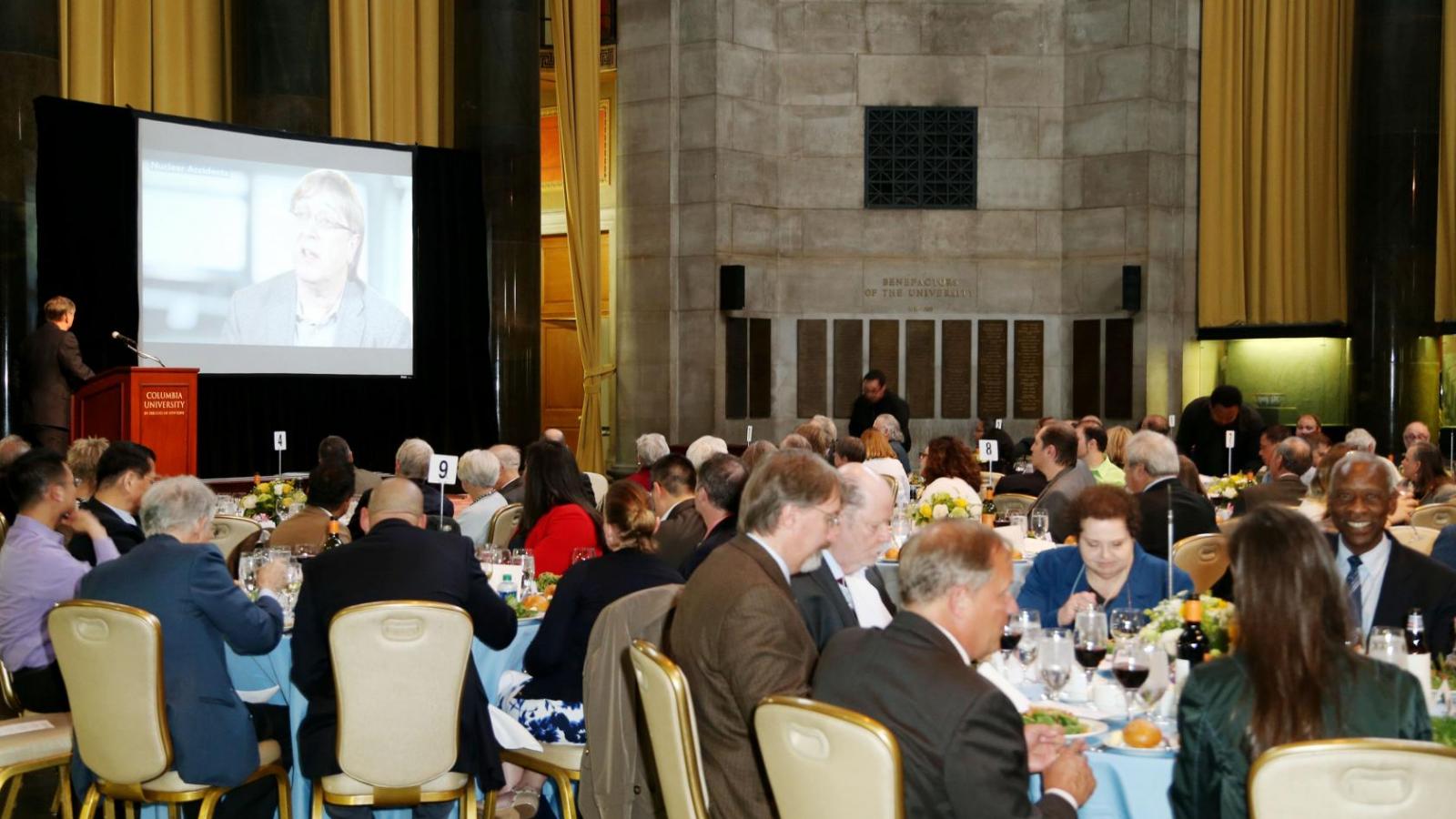 Large group of people seated at dinner tables with blue table cloths in a large room with stone walls and projection screen with podium towards the front of the room