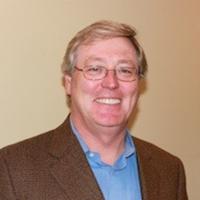 man with brown suit coat, blue dress shirt and glasses smiling for headshot