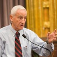 Man with gray hair in light colored dress shirt, red and black tie at podium giving a speech