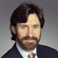 Man with dark hair and beard wearing blue suit jacket, white dress shirt, and a tie posing for a head shot