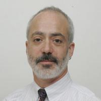 Man with greying hair and beard, wearing white pin striped dress shirt and tie posing for headshot