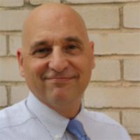 Man in striped light colored dress shirt and light blue tie posing for headshot with brick wall in background